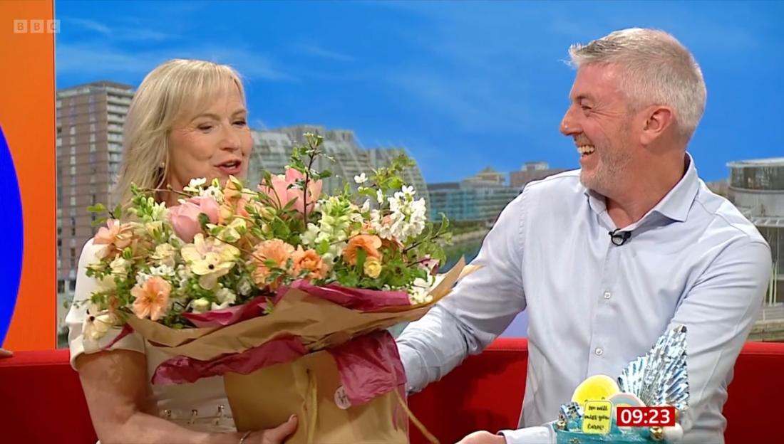 A blonde woman holding a large bouquet of flowers looks at a laughing man in a light blue shirt.