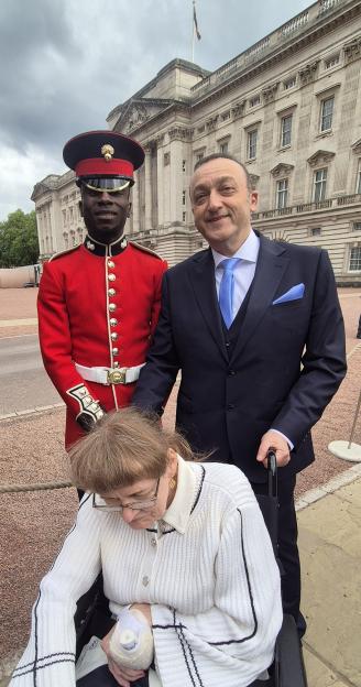 John Manitara, a man in a suit, with his wife Gillian, who is in a wheelchair and appears to be sleeping, standing next to a royal guard in a red uniform, with a building in the background.
