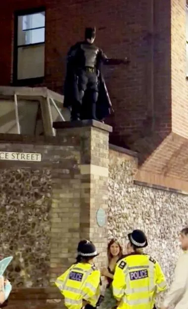 A person in a Batman costume stands on a brick wall above two police officers and two women on a street at night.