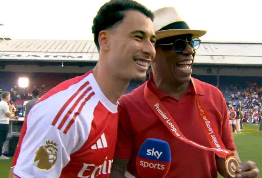 A soccer player in a red and white jersey smiles next to an older man wearing a hat, sunglasses, and a red shirt with a "Premier League" medal around his neck.
