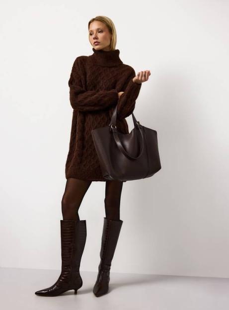 Woman wearing a brown jumper dress, tights, knee-high crocodile boots, and holding a brown tote bag.