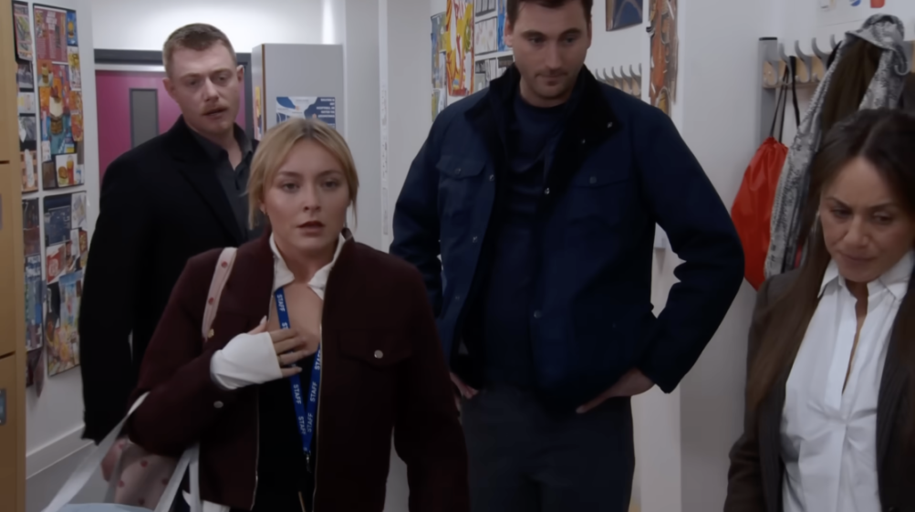 A young woman with a white cast on her left hand and a blue "STAFF" lanyard around her neck, stands in a hallway surrounded by two men and a woman.