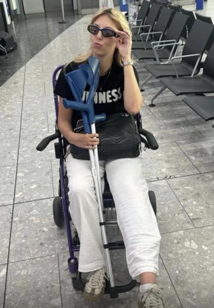 A woman in a wheelchair holding crutches, wearing a black t-shirt, white pants, and sunglasses, pouting at the camera in an airport.