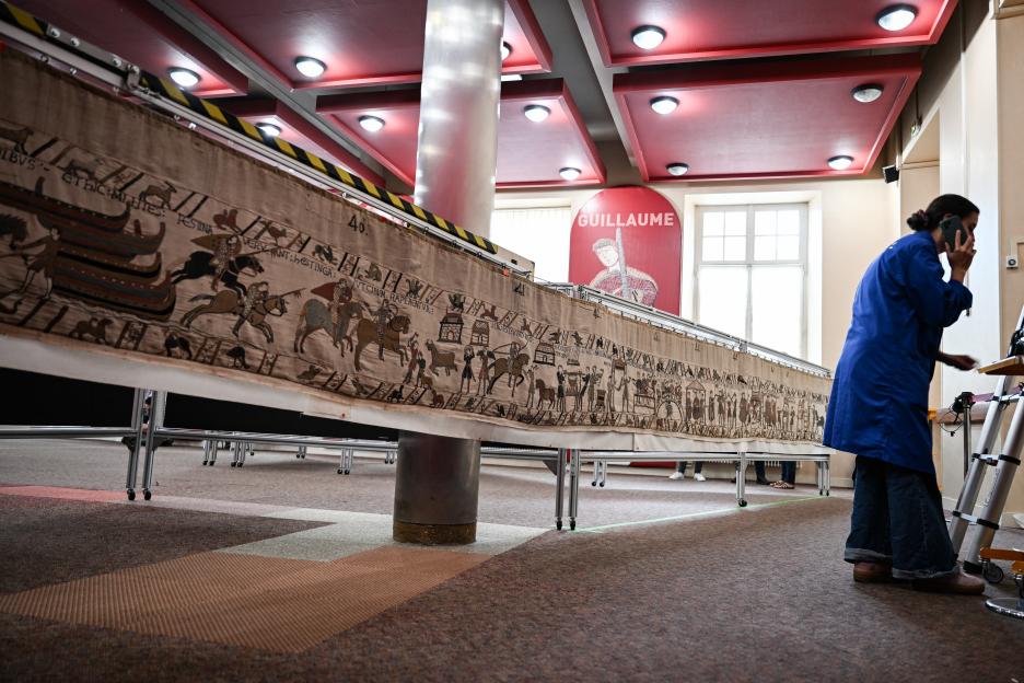 A worker on a phone prepares the Bayeux Tapestry for packing and transfer, in a room with a large red "GUILLAUME" sign.