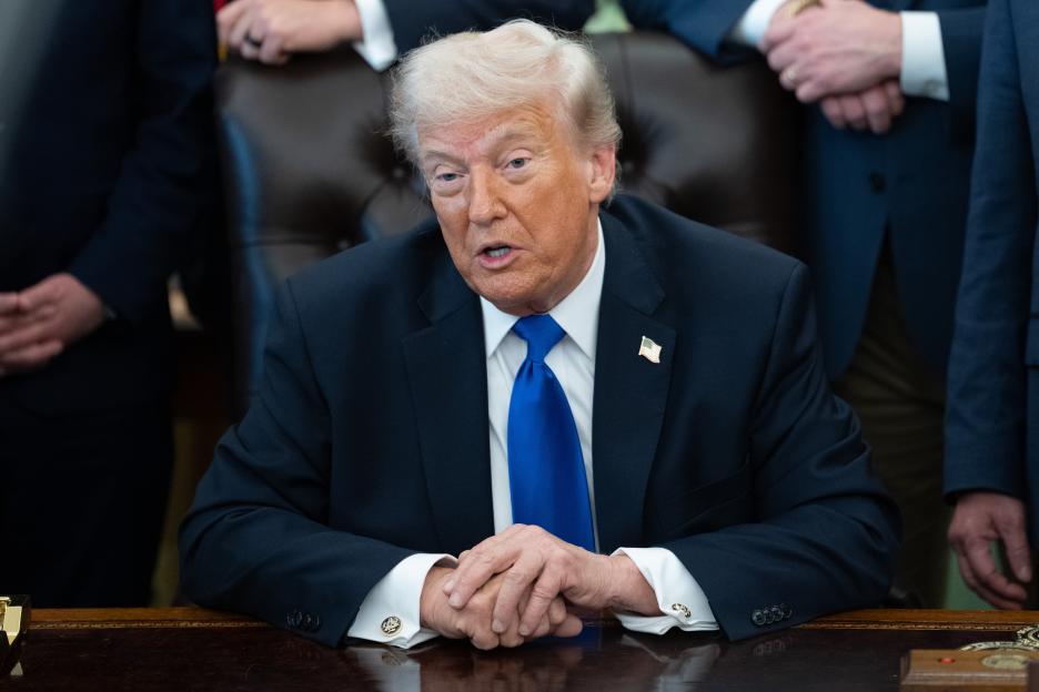 Donald Trump in a dark suit and blue tie speaking at a desk in the Oval Office.