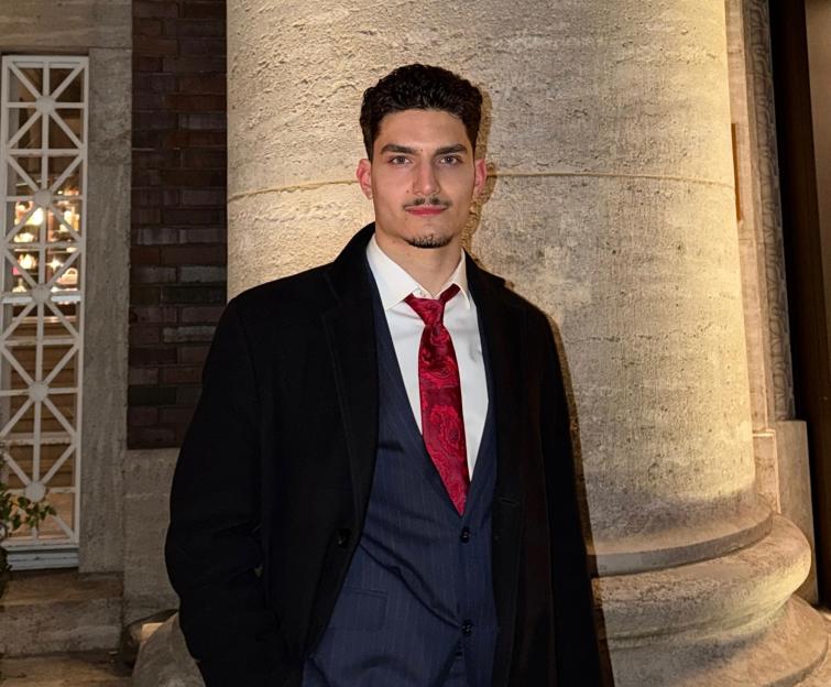 A man in a black coat, blue pinstriped vest, white shirt, and red tie stands next to a large stone column.