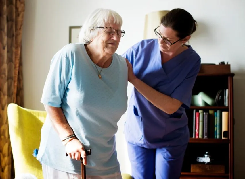 A nurse in blue scrubs helps an elderly woman with a cane to stand.