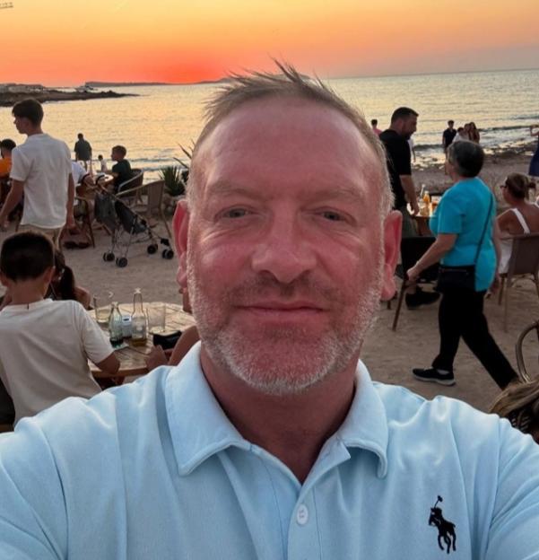 A man in a light blue polo shirt takes a selfie with a sunset over the ocean in the background.