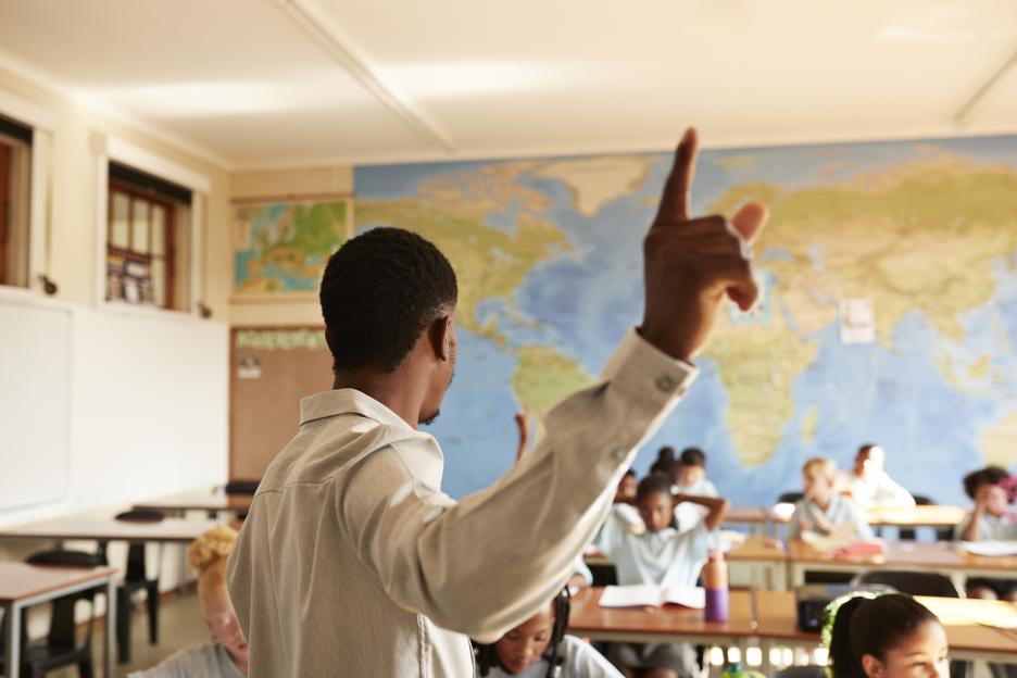A young teacher points to a map while teaching a diverse group of elementary school students.