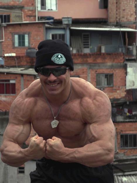A muscular man wearing a beanie and sunglasses flexing with a favela in the background.