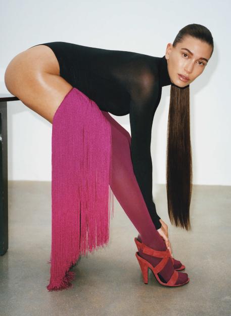 Hailey Bieber models a sheer black bodysuit and a long pink fringed skirt, with brown hair draped to the floor, wearing maroon heels.