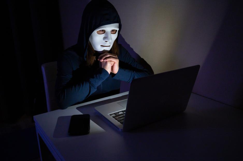 A person wearing a white mask and a dark hoodie, with hands clasped, sits in front of a laptop in a dimly lit room.