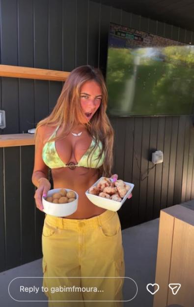 A woman in a bikini top and yellow pants winks at the camera while holding two bowls of snacks, with a soccer game playing on a TV in the background.