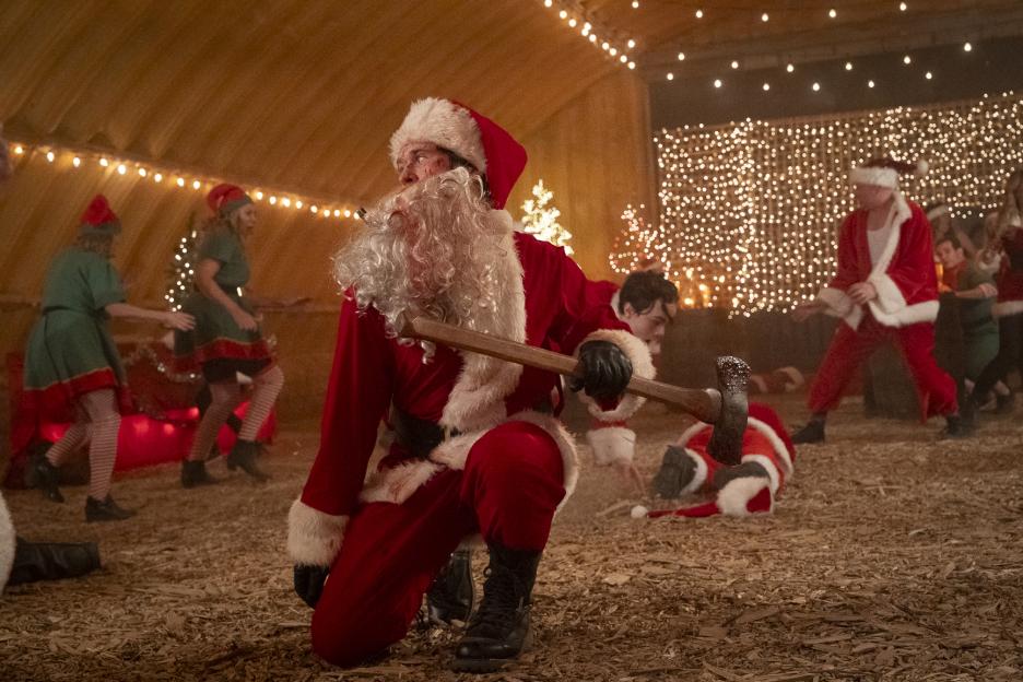 Rohan Campbell as Billy Chapman, dressed as Santa Claus and holding an axe, surrounded by people in festive costumes in a Christmas-decorated scene.