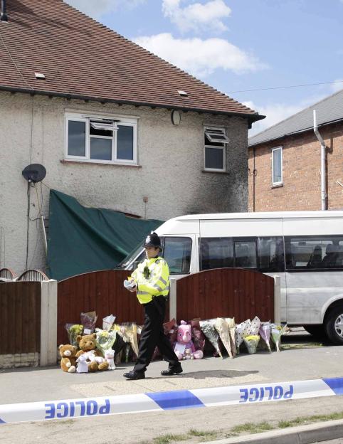A police officer walks past a fire-damaged house with floral tributes and stuffed animals laid outside, marked by police tape.