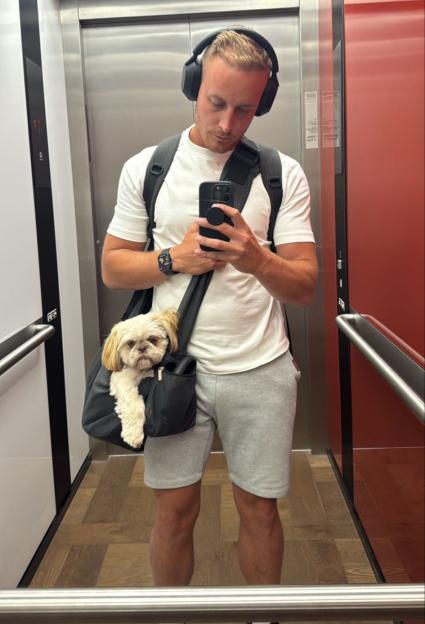 A man in an elevator taking a mirror selfie with his dog in a carrier.