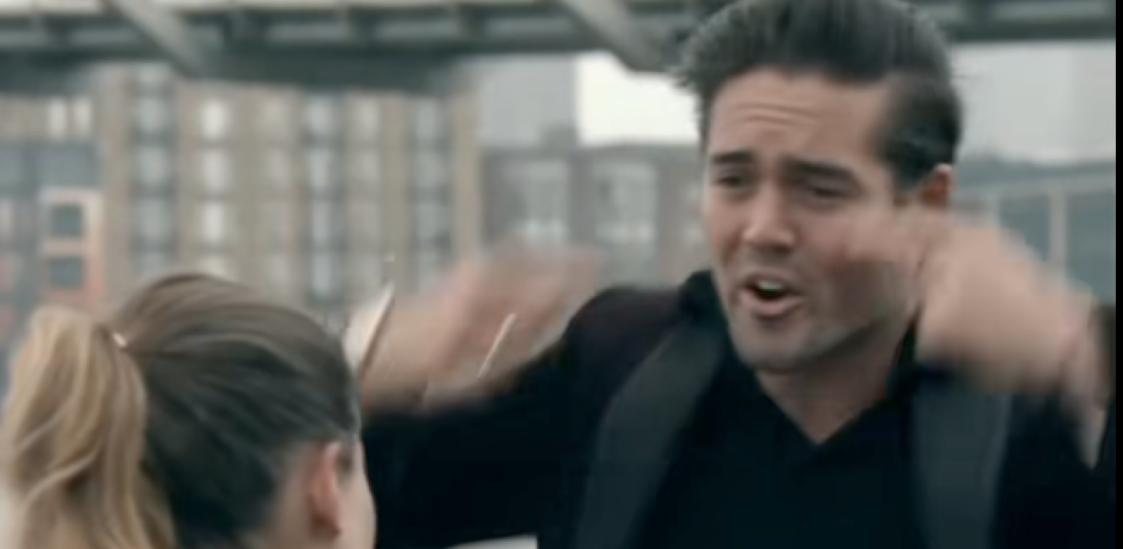 Man in black suit shouting with hands raised, facing a woman.