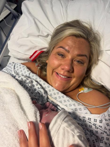 A woman in a hospital gown smiles at the camera while holding her newborn baby's foot.