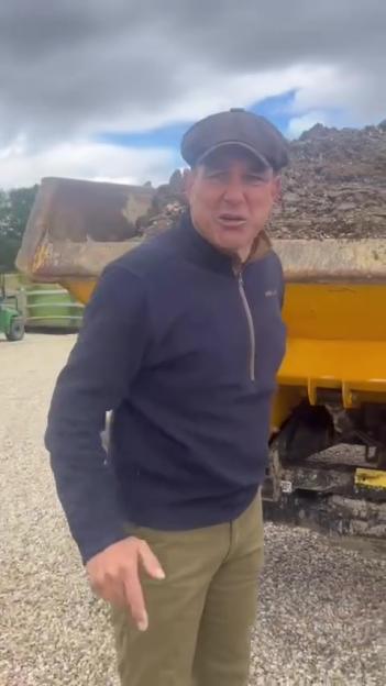 Vinnie Jones in a navy pullover and brown flat cap standing in front of a yellow dump truck.
