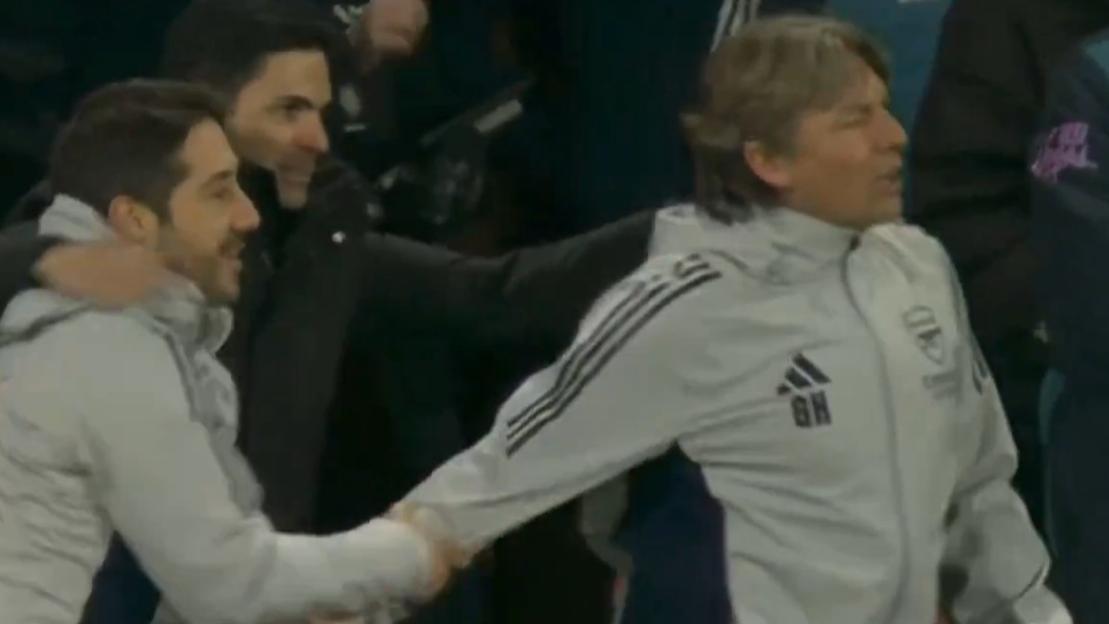 Three men on the sidelines of a soccer match, one holding another's hand.
