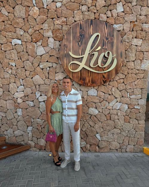 Leandro Trossard and Laura Hilven standing in front of a stone wall with a wooden "Lío" sign.