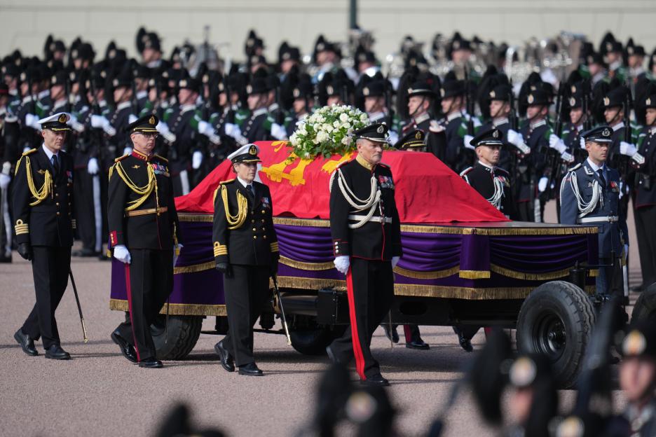 The coffin of King Harald V of Norway, covered in a red and gold flag and adorned with white flowers, is transported on a wheeled carriage by military personnel in uniform.