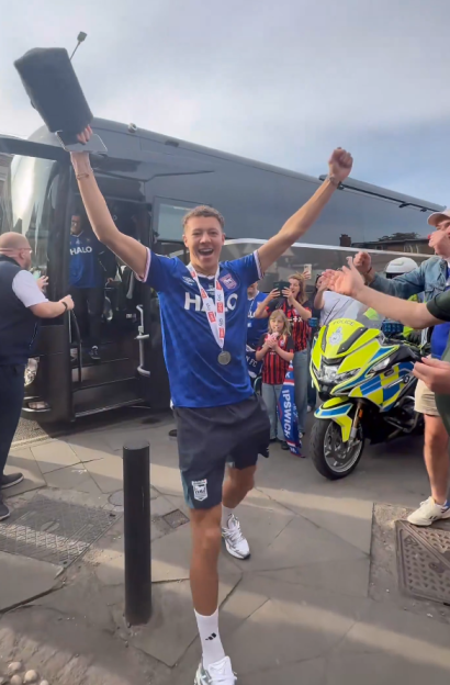 A male soccer player in a blue uniform with medals celebrates with arms raised next to a team bus and police motorcycle.