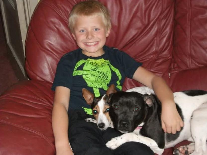 Andre Fairbairn smiling on a red couch, hugging two dogs.
