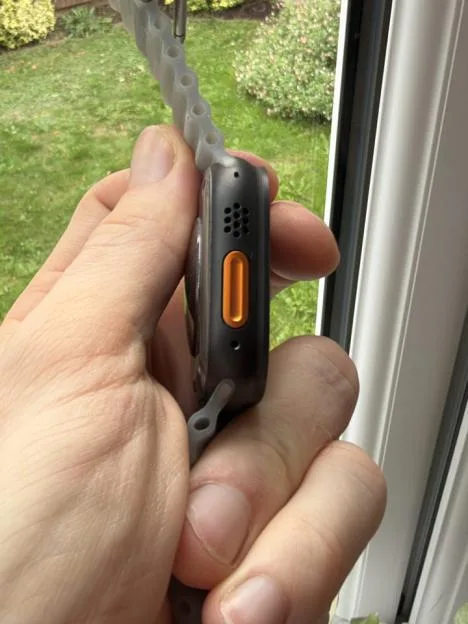 Close-up of a hand holding the left side of an Apple Watch Ultra, showing the orange Action button, speaker grille, and part of the Ocean Band.