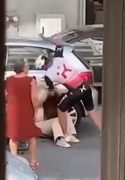 An elderly woman in a red dress hitting a man who is kneeling by a car.
