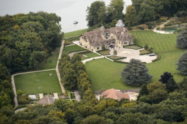 Aerial view of a sprawling estate with a large house, green lawns, gardens, and a soccer field next to a body of water.