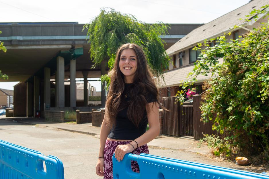 A young woman named Leilea Jammali smiles, standing with a blue barrier in the foreground, with residential buildings and a motorway overpass in the background.