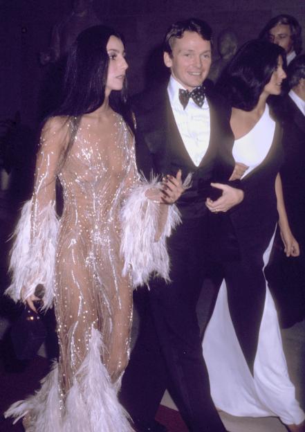 Cher in a sheer, embellished gown with feathers, accompanied by Bob Mackie in a tuxedo and another woman in a black and white outfit.
