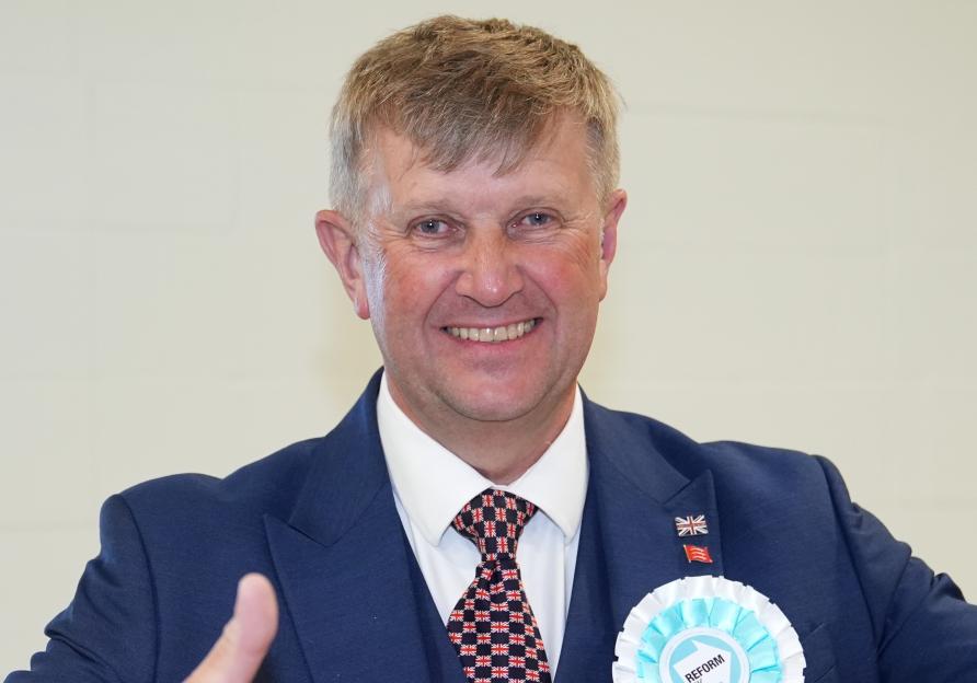 A smiling man in a suit, with a Reform UK rosette and Union Jack tie, gives two thumbs up.