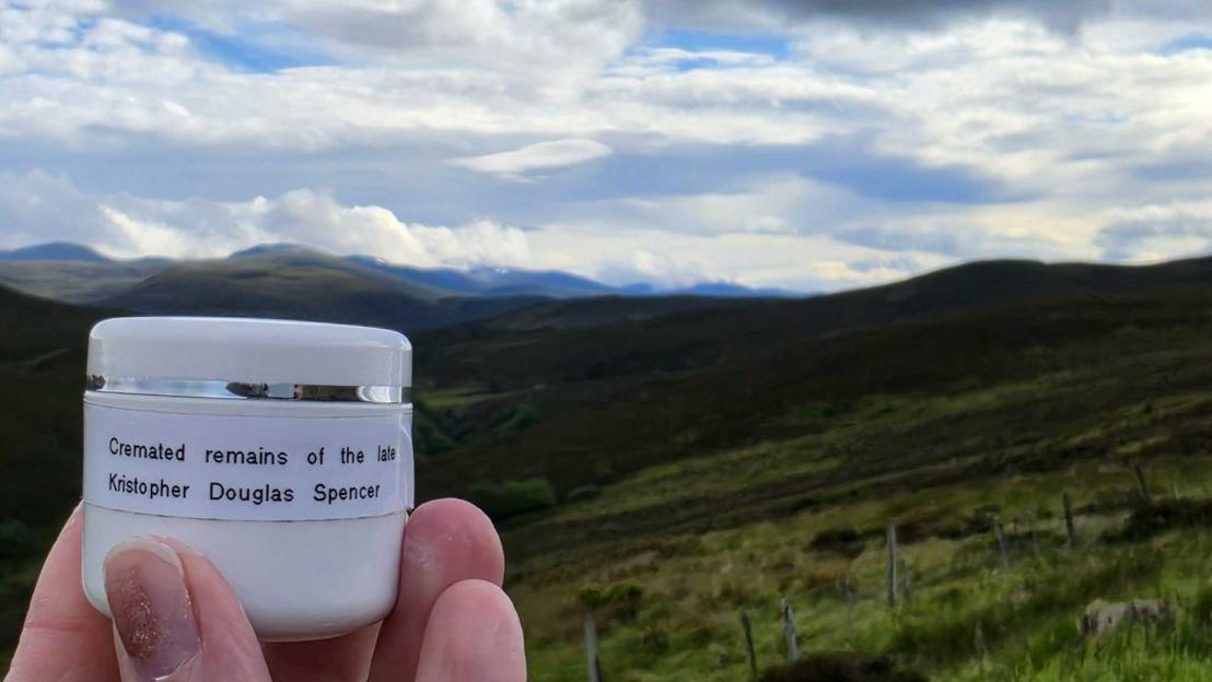 A person holding a small white container labeled "Cremated remains of the late Kristopher Douglas Spencer" in front of a mountainous landscape.