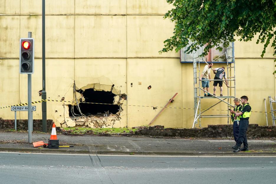 Police and workers assess the damage to a building with a large hole in its wall, cordoned off by yellow tape.