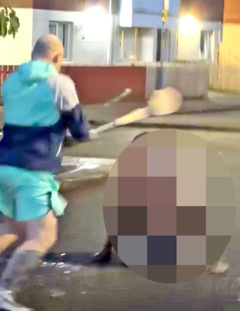 A man in a blue and white jacket and shorts, hitting another person, whose body is blurred, with a large wooden paddle.