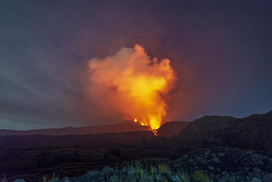 Mount Etna erupting at night with bright orange smoke against a starry sky.
