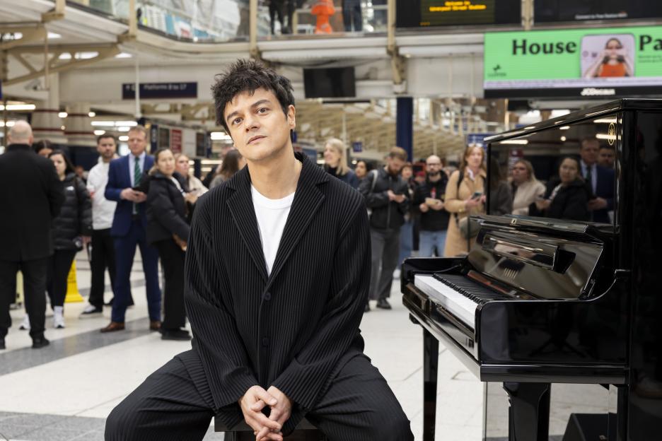 Jamie Cullum sitting next to a black Yamaha piano in a busy train station.