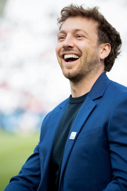 Sam Thompson laughing while wearing a blue suit jacket and a Soccer Aid for UNICEF pin.