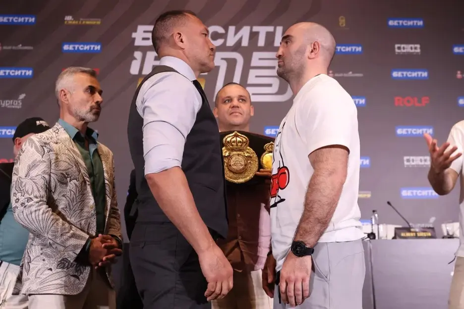 Two male boxers face off at a weigh-in with a championship belt visible between them.
