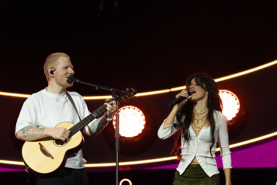 Ed Sheeran playing guitar and Camila Cabello singing into a microphone on stage.