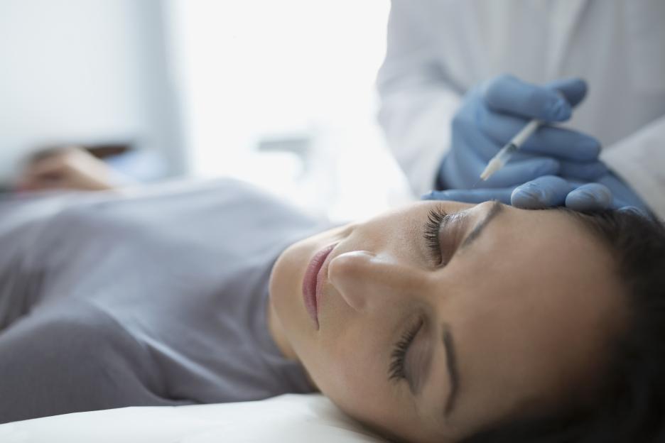 A woman lies with her eyes closed, receiving a Botox injection in her forehead from a gloved hand holding a syringe.