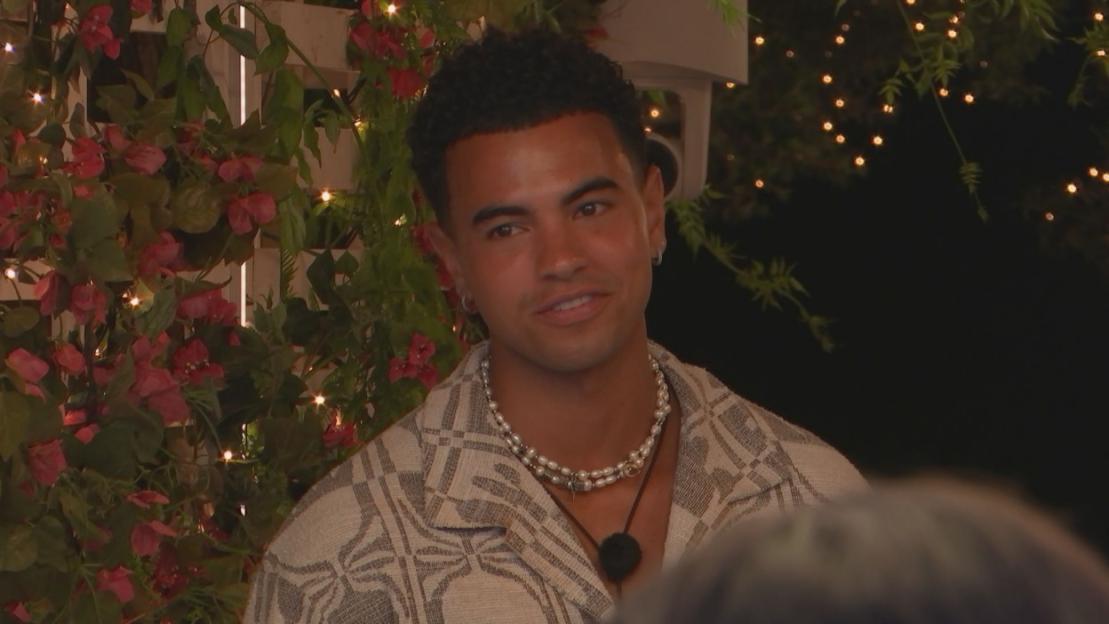 A Love Island contestant, a young man, in a patterned shirt and pearl necklace, smiles against a backdrop of foliage and fairy lights.