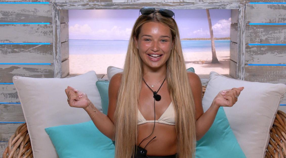 Lucinda Strafford of Love Island smiles while seated on a wicker chair with an ocean view in the background.