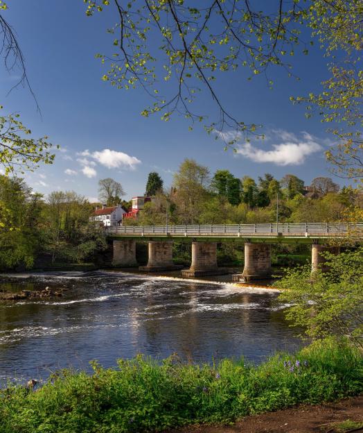 A concrete and stone bridge spanning a river, with trees and houses on the far bank.