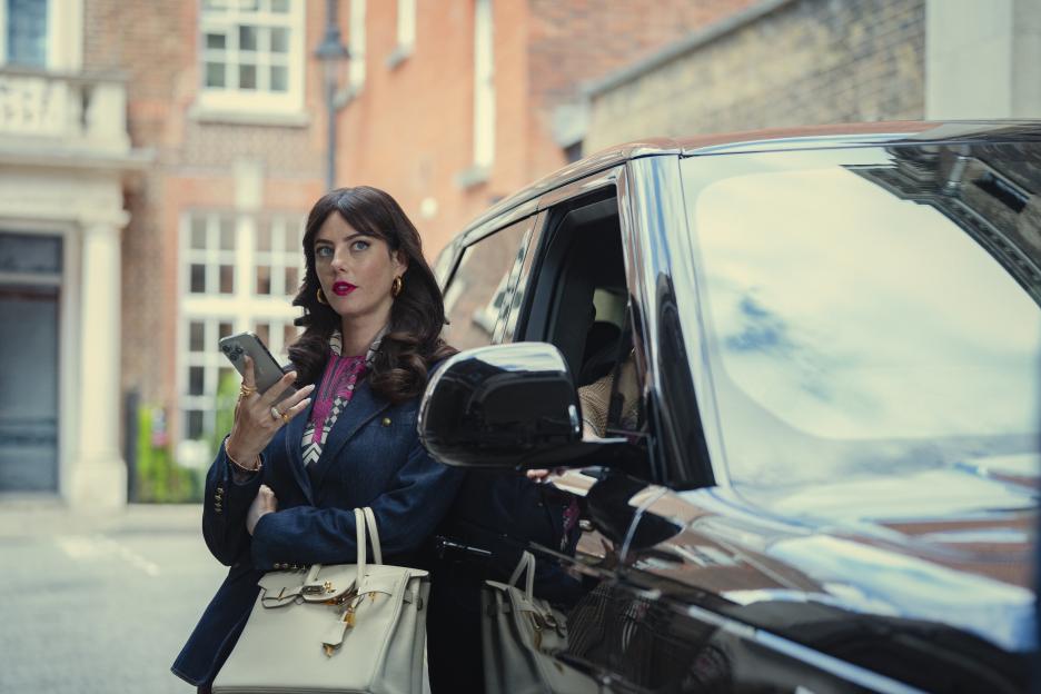 Kaya Scodelario as Susie Glass in The Gentlemen Series 2, standing next to a black car, holding a phone and a cream handbag.