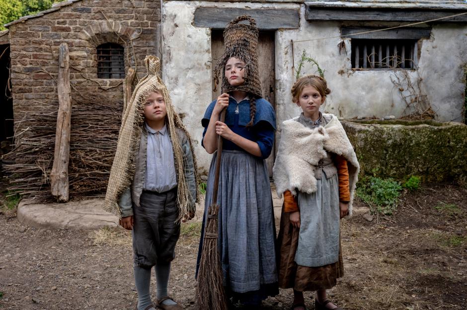 Three children in historical clothing and hats made of straw, standing in front of a stone building.