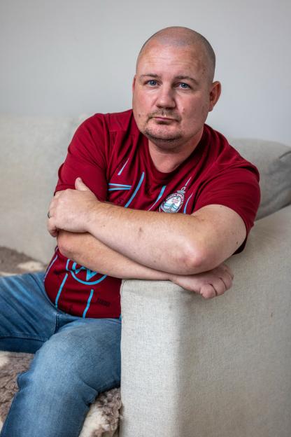 Gareth Burns, diagnosed with silicosis, seated on a couch with arms crossed, wearing a maroon and blue shirt.
