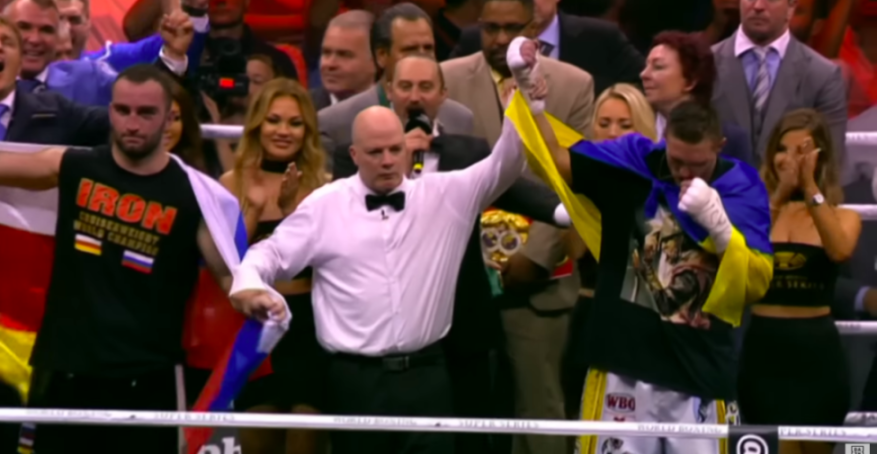 A boxing referee raises the arms of two boxers, one with a Russian flag and the other with a Ukrainian flag, in a boxing ring.